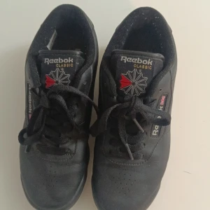 Svarta Reebok Classic sneakers - Klassiska svarta Reebok sneakers i skinn med låg profil och snörning. Ikonisk logga på tungan och sidan, samt diskreta perforeringar framtill för extra ventilation. Perfekta för en clean och tidlös streetstil.