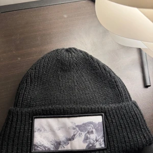 Svart ribbad mössa med tryck - Snygg svart ribbad mössa med ett unikt tryck på framsidan. Perfekt för att hålla värmen under kyliga dagar. Stilren och enkel design som passar till det mesta. Från revenite