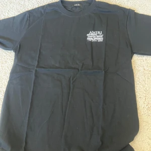 Svart t-shirt från Amiri - Snygg svart t-shirt från Amiri med tryck på både fram- och baksidan. Framsidan har en liten vit text på bröstet medan baksidan har ett stort tryck med text och rosa benkors. Perfekt för en cool och avslappnad stil.