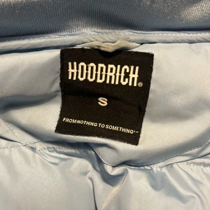 Ljusblå pufferjacka från Hoodrich - Säljer en ljusblå pufferjacka från Hoodrich med dragkedja och textdetaljer på bröstet. Jackan har en stor huva och är perfekt för kyligare dagar. Den är långärmad och har en modern streetwear-look.