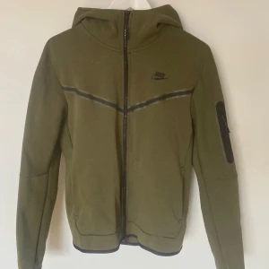 Olivgrön Nike tech  - Säljer en olivgrön Nike Tech jacka med huva från Nike. Jackan har en dragkedja framtill och en ficka med dragkedja på ärmen.