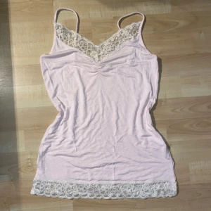Ljusrosa linne med spetsdetaljer - Säljer ett ljusrosa linne med vackra spetsdetaljer vid halsringningen och nederkanten. Linnet har smala axelband och en mjuk, bekväm passform. Perfekt för varma sommardagar eller som ett lager under andra plagg.