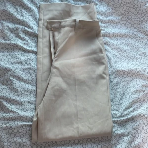 Beige kostymbyxor från NA-KD - Snygga beige kostymbyxor från NA-KD med hög midja och stilren design. Byxorna har en dragkedja framtill och hällor för bälte. Perfekta för en chic och elegant look.