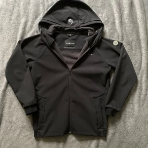 Svart jacka från Sail Racing - Snygg svart jacka från Sail Racing med huva och dragkedja. Jackan har en stilren design med logotypdetaljer på bröstet och ärmen. Jackan är i tunt material. Storlek 176. Vid frågor eller funderingar skicka ett dm💫