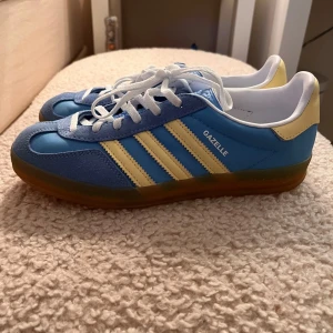 Adidas Gazelle i blått och gult - Säljer ett par snygga Adidas Gazelle sneakers i blått och gult. Skorna är i mycket bra skick och har en bekväm passform. Perfekta för både vardag och fest!
