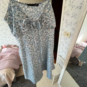 Blommig blå klänning med knytband - Charmig blå klänning med blommigt mönster och knytband framtill. Perfekt för en somrig look. Klänningen har en avslappnad passform och ärmlös design.