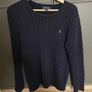 Mörkblå stickad tröja från Polo Ralph Lauren - Säljer en snygg mörkblå stickad tröja från Polo Ralph Lauren. Tröjan har ett klassiskt kabelstickat mönster och en liten grön logga på bröstet. Perfekt för kyligare dagar med sin långärmade design och ribbade kanter.