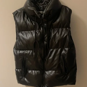 Svart dunväst från New Roni - Snygg svart dunväst från New Roni med dragkedja och hög krage. Perfekt för kyliga dagar när du vill hålla värmen utan att kompromissa med stilen. Västen har praktiska fickor med dragkedja för säker förvaring.