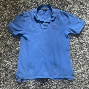 Blå pikétröja från Polo Ralph Lauren - Säljer en klassisk blå pikétröja från Polo Ralph Lauren med korta ärmar och en broderad röd logga på bröstet. Perfekt för en stilren och avslappnad look. 💙