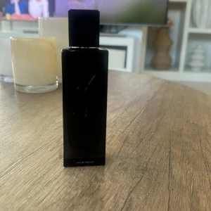 Svart parfymflaska från Yves Saint Laurent - Elegant svart parfymflaska från Yves Saint Laurent med en stilren design. Flaskan har en fyrkantig form och pryds av märkets ikoniska logotyp i relief. Perfekt för den som söker en sofistikerad doftupplevelse.