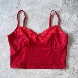 Röd spetsdetaljerad linnetopp - Snygg röd linnetopp med spetsdetaljer och justerbara axelband. Toppen har en söt rosett framtill och är perfekt för en trendig look.