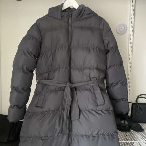 Svart dunjacka från Threadbare - Snygg svart dunjacka från Threadbare med huva och bälte i midjan. Jackan har en dragkedja framtill och långa ärmar. Perfekt för kalla dagar när du vill hålla dig varm och stilren. Köpt från asos för 625kr och oanvänd.