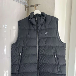 Svart dunväst från GANT - Snygg svart dunväst från GANT med dragkedja och hög krage. Perfekt för lager-på-lager under kyliga dagar. Västen har två praktiska fickor med dragkedja framtill. Ny pris 2100 mitt pris 500