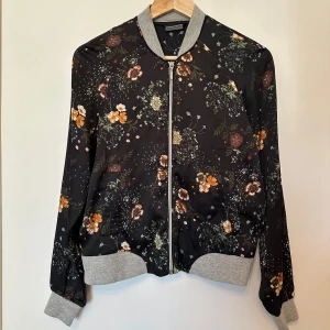 Blommig bomberjacka från Vero Moda - Snygg bomberjacka från Vero Moda med blommigt mönster i orange, grönt och vitt på en svart bakgrund.