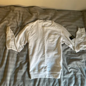 Vit långärmad tröja från Stone Island Junior - Säljer en stilren vit långärmad tröja från Stone Island Junior. Tröjan har en bröstficka och det ikoniska Stone Island-märket på ärmen. Perfekt för en avslappnad och trendig look. Model 179cm och 65kg. Nypris 1660kr