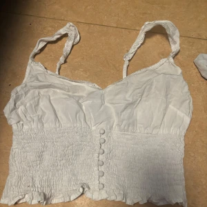 Vit linnetopp från NA-KD - Säljer en söt vit linnetopp från NA-KD med smockdetaljer och knappar framtill. Toppen har justerbara axelband med spetsdetaljer och en croppad passform. Perfekt för varma dagar!