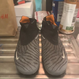 Nike fotbollsskor med zebramönster - Snygga fotbollsskor från Nike med zebramönster i svart och grått. Skorna har en rund tå och är designade för optimal prestanda på planen. De har en strumpliknande passform och en iögonfallande orange detalj på insidan.