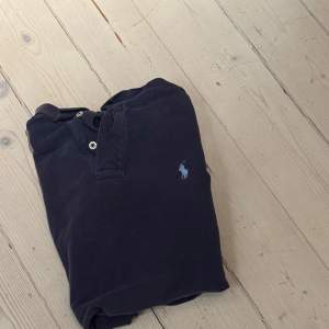 Säljer en klassisk mörkblå pikétröja från Ralph Lauren med korta ärmar och knappar vid halsen. Den har den ikoniska loggan broderad på bröstet. Perfekt för en stilren look!
