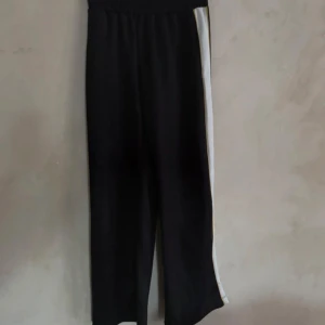Svarta tracksuits - Snygga svarta byxor med vita ränder längs sidorna. De har en elastisk midja för extra komfort och en avslappnad passform. Perfekta för en sportig look eller en avslappnad dag.
