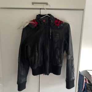 Hugo boss  skinnjacka med röd och vit pälsdetalj - Unik svart skinnjacka från Hugo Boss. Den har ett rött och  svart mönster på insidan av luvan i tunn päls, och vit päls på luvan - vilken kan bli som en krage om man bara drar ner dragkedjan. Jackan har även en liten mobilväska på insidan, kors på båda ärmarna och lite nitar. 