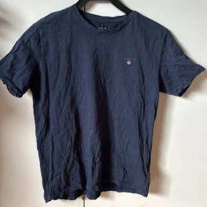 Mörkblå t-shirt från GANT - Säljer en stilren mörkblå t-shirt från GANT med en liten broderad logga på bröstet. T-shirten är kortärmad och gjord i mjukt bomullsmaterial. Perfekt för en avslappnad look.