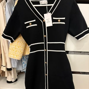 Svart klänning med vita detaljer  - Snygg svart klänning med vita kontrastdetaljer och guldknappar från Paper Moon. Klänningen har korta ärmar och en v-ringad design med dekorativa fickor framtill. Perfekt för en stilren look! Aldrig använd. 
