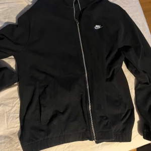 Svart hoodie från Nike - Säljer en svart hoodie från Nike med dragkedja och huva. Jackan har långa ärmar och en liten Nike-logga på bröstet. Perfekt för en avslappnad stil.