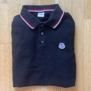 Svart pikétröja från Moncler - Snygg svart pikétröja från Moncler med klassisk krage och knappar framtill. Tröjan har röda och vita detaljer på krage och ärmslut samt Monclers logga på bröstet. Perfekt för en stilren look.