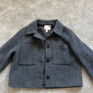 Grå ulljacka från H&M - Säljer en stilren grå ulljacka från H&M. Jackan har en klassisk krage och knäppning med knappar framtill. Den har två stora fickor på framsidan och är perfekt för kyligare dagar. Passar bra till både vardag och mer uppklädda tillfällen💕