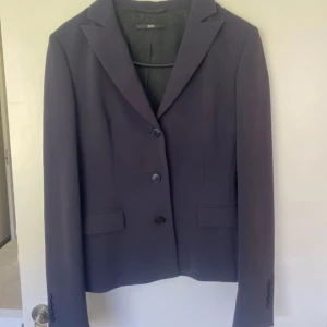 Mörkblå kavaj från Hugo Boss - Elegant mörkblå kavaj från Hugo Boss med klassisk design och knappar framtill. Perfekt för en stilren look. Kavajbyxorna kommer även med alltså är det ett helt sett. Settet kan vara liten i storleken så det passar troligen dig som har xs i storlek. Det är inga fickor där det ser ut att vara det utan det är bara en design. Den är i skick som ny och nästan aldrig använd. 