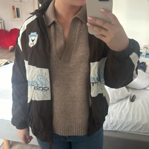 Brun och vit vindjacka med Monaco-märke - Säljer en brun och vit vindjacka med coola detaljer som ett Monaco-märke på bröstet och stora siffror på ryggen. Jackan har dragkedja och långa ärmar, perfekt för en sportig look. Den har en avslappnad passform och är idealisk för vår och höst.