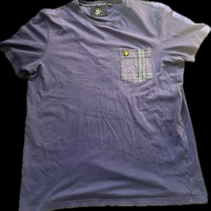 Lila/blå t-shirt från Lyle & Scott - Snygg blå t-shirt från Lyle & Scott med en rutig bröstficka och deras ikoniska logga. Perfekt för en avslappnad stil. T-shirten är kortärmad och gjord i mjukt material.