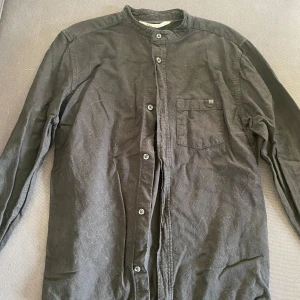 Svart skjorta från H&M barn  - Snygg svart skjorta från H&M med ståkrage och knappar framtill. Skjortan har en bröstficka och långa ärmar, perfekt för en stilren look.