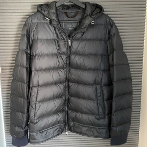 Svart pufferjacka från Massimo Dutti - Stilren pufferjacka från Massimo Dutti. Jackan är i mörkgrå/svart med en marinblå ”resår” baktill. Perfekt för kyliga dagar med sin quiltade design som håller värmen. Jackan har två fickor på sidorna samt en innerficka.