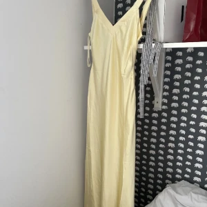 Ljusgul långklänning med v-ringning - Säljer en elegant ljusgul långklänning med v-ringning och korsade band i ryggen. Perfekt för sommarens festligheter. Klänningen har en stilren design som ger en sofistikerad look. Passar utmärkt för både formella och avslappnade tillfällen.