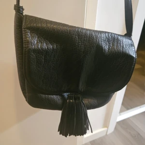 Svart axelväska med fransdetalj - Snygg svart axelväska i skinn med en stilren fransdetalj på framsidan. Väskan har en praktisk dragkedja upptill och en justerbar axelrem. Perfekt för att bära dina nödvändigheter med stil.