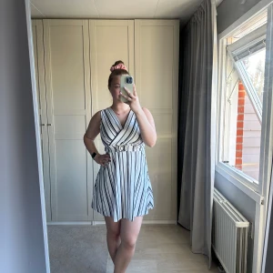 Randig klänning från SisterS Point - Snygg ärmlös klänning från SisterS Point i vitt med svarta ränder. Klänningen har en omlott-design och ett bälte i midjan med dekorativt spänne. Perfekt för en stilren look!