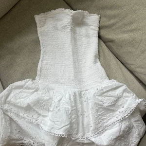 Vit axelbandslös klänning från Ginatricot - Säljer en söt vit axelbandslös klänning från Ginatricot. Klänningen har en smockad överdel och en volangkjol med broderade detaljer. Perfekt för sommarens alla tillfällen! 🌸