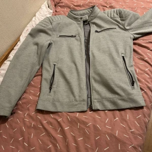 Grå jacka med dragkedjor - Snygg grå jacka med dragkedjor på bröstet och sidorna. Jackan har en modern design med långa ärmar och en stilren look. Perfekt för både vardag och lite finare tillfällen. Pris kan diskuteras 