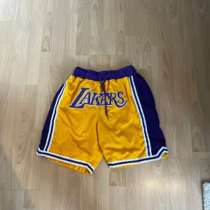 Gula Lakers shorts - Snygga gula Lakers shorts med lila och vita detaljer. De har snörning i midjan och är perfekta för sportiga aktiviteter. Lakers-loggan är tryckt på framsidan.
