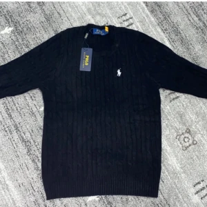 Svart och vit kabelstickad tröja Polo - Två kabelstickade tröjor från Polo Ralph Lauren, en i svart och en i vit. Båda har rund hals, lång ärm och den klassiska broderade loggan på bröstet. Tröjorna är gjorda i mjuk bomull och har ribbade muddar vid ärmslut och nederkant. Perfekt för dig som gillar stilren och tidlös design.