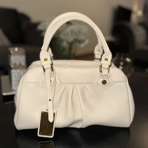 Vit handväska från Marc Jacobs - Snygg vit handväska från Marc Jacobs i mjukt skinn med guldfärgade metalldetaljer och dubbla handtag. Väskan har en klassisk form, dekorativa veck framtill och en metallbricka med logga. Perfekt för dig som vill ha en stilren och lyxig accessoar.