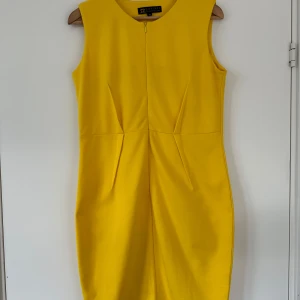 Gul ärmlös kortklänning Shez XL - Snygg gul kortklänning från Shez i storlek XL. Klänningen är ärmlös med rund halsringning och har en dold dragkedja framtill. Materialet är mjukt och känns som bomull eller polyester. Snygga sömdetaljer framtill ger en cool och modern look.