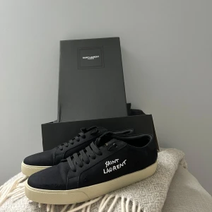 Svarta Saint Laurent sneakers - Snygga svarta sneakers från Saint Laurent med vit broderad logga på sidan och kontrasterande vit sula. Skorna har rund tå, platt sula och klassisk snörning. Materialet är canvas med detaljer i skinn, vilket ger en stilren och modern vibe.