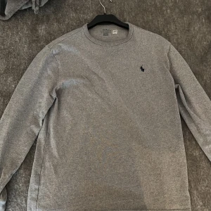 Grå långärmad tröja från Polo Ralph Lauren - Säljer en stilren grå långärmad tröja från Polo Ralph Lauren med klassisk rund hals och den ikoniska lilla logon broderad på bröstet. Tröjan är i mjuk bomull och har en normal passform, perfekt för chill dagar eller när du vill hålla det enkelt.