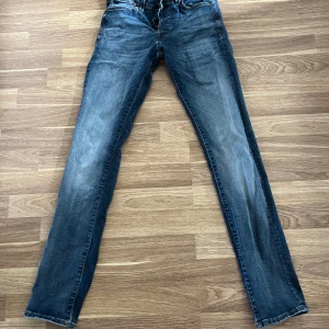 Slim fit jeans från Jack & Jones - Snygga blå slim fit jeans från Jack & Jones, modell Glenn. Jeansen har klassisk femficksdesign, smal passform och coola slitningar framtill. Tillverkade i mjukt denim med orangea sömmar och metallknappar. Perfekta för dig som gillar en modern och stilren look.
