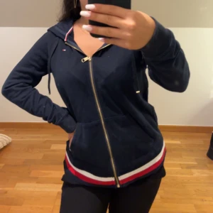 Marinblå hoodie från Tommy Hilfiger - Snygg marinblå hoodie från Tommy Hilfiger med dragkedja i guld och huva med snörning. Tröjan har långa ärmar och ribbad kant med röd, vit och marinblå rand längst ner. Perfekt för chill dagar och har klassiska Hilfiger-detaljer vid halsen. Nästan aldrig använd men har en liten fläck som förmodligen går bort i tvätten, bilder på logo osv kan skickas!