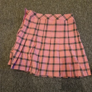 Rutig rosa plisserad kortkjol - Säljer en superfin rosa kortkjol med svart och vit rutigt mönster. Kjolen är plisserad och har en klassisk skolstil som är perfekt för att skapa en trendig look. Passar dig som gillar färg och mönster. Dragkedja i sidan för enkel påklädning.