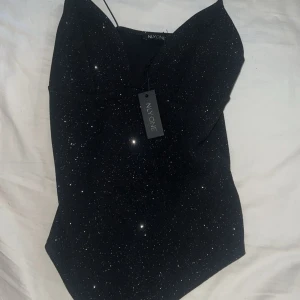 Svart glittrig body från nly  - Supersnygg svart body från NLY One med tunna axelband och glittrigt material som verkligen poppar. Bodyn  har en v-ringad front och figurnära passform som ger en chic siluett. Perfekt för dig som vill sticka ut med lite extra glitter.