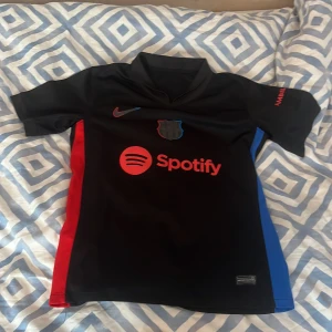 FC Barcelona Gavi #6 Nike fotbollströja - Snygg svart FC Barcelona fotbollströja med Gavi #6 på ryggen, tillverkad av Nike. Tröjan har röda och blå detaljer på sidorna, röd Nike-logga och Spotify-tryck på bröstet. Kortärmad med krage och tryck på ärmen. Perfekt för dig som älskar Barca och Gavi.